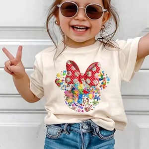 Peut inclure: Un t-shirt crème avec un motif Minnie Mouse. Le motif comprend un nœud rouge à pois et un collage de formes et de motifs colorés, notamment des cœurs, des étoiles et des pièces de puzzle. Le t-shirt est associé à un jean en denim bleu.
