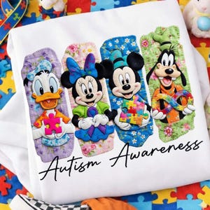 Può includere: Felpa bianca con Paperino, Minnie, Topolino e Pippo, ognuno con un pezzo di puzzle. La scritta "Autism Awareness" è in corsivo. I personaggi sono su sfondi colorati.