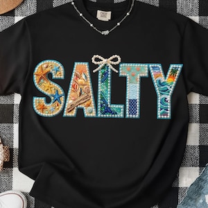 May include: Black t-shirt with the word "SALTY" in a colorful, beach-themed design. The letters are filled with seashells, starfish, and ocean waves. A bow is above the "T". A silver necklace is visible.