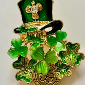 May include: A St. Patrick's Day brooch featuring a green top hat with gold trim and a clear gem. The brooch also includes green shamrocks and a gold coin with a shamrock design. The brooch is a festive accessory.