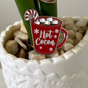 May include: A red enamel pin shaped like a mug of hot cocoa with marshmallows and a candy cane. The mug has a silver rim and handle and says "Hot Cocoa" with snowflakes.