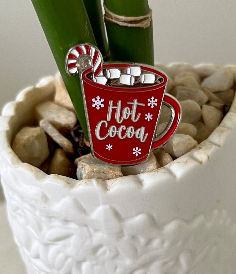 May include: A red enamel pin shaped like a mug of hot cocoa with marshmallows and a candy cane. The mug has a silver rim and handle and says "Hot Cocoa" with snowflakes.