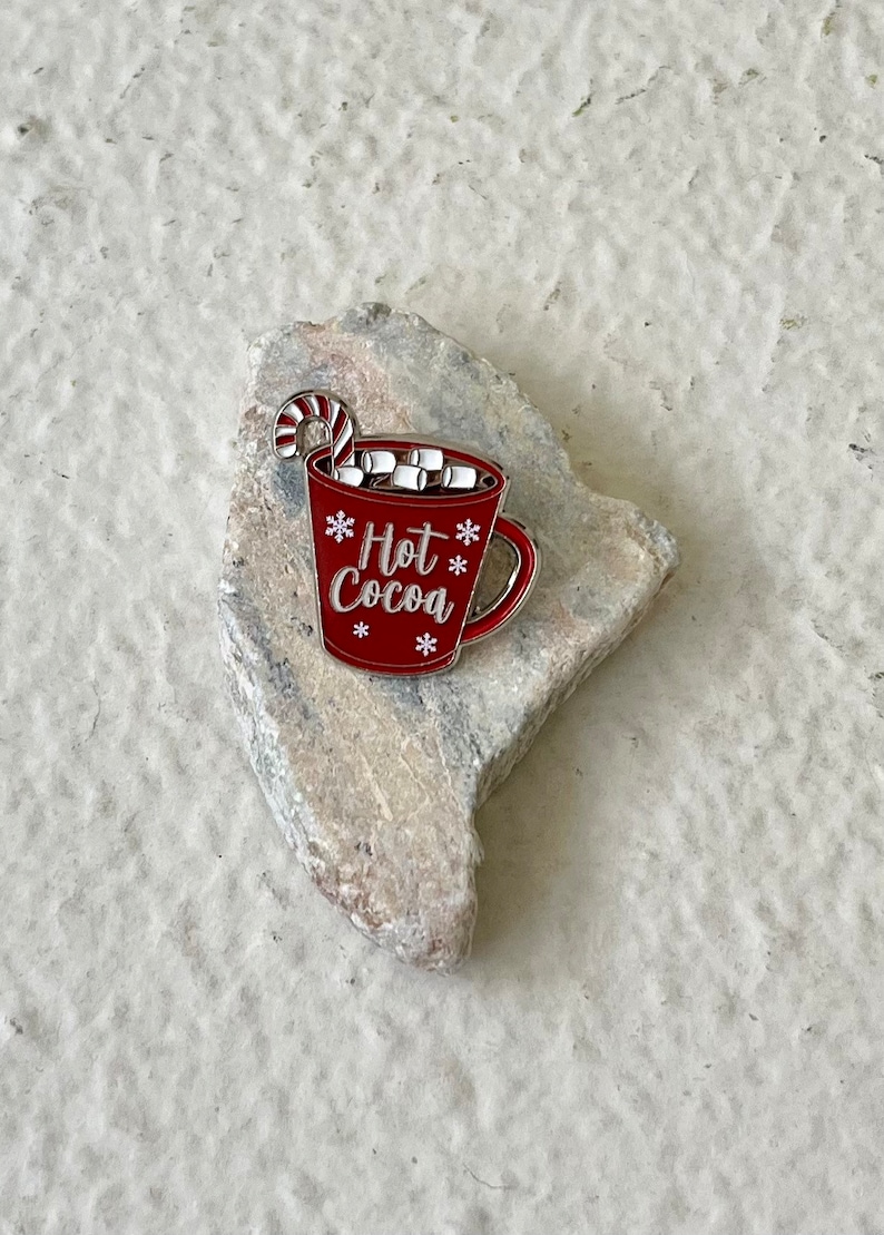 May include: Red enamel pin of a mug with white marshmallows and a candy cane, with the text "Hot Cocoa" in white lettering and snowflakes.