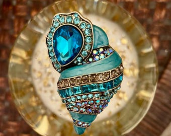 May include: A decorative seashell-shaped brooch with a teal and gold color scheme. The brooch is adorned with various sizes of blue and iridescent crystals. The top of the brooch features a large, teardrop-shaped blue crystal.