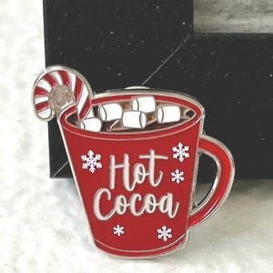 May include: Red enamel pin shaped like a mug of hot cocoa with marshmallows and a candy cane. The mug has white snowflakes and the text "Hot Cocoa" on it.