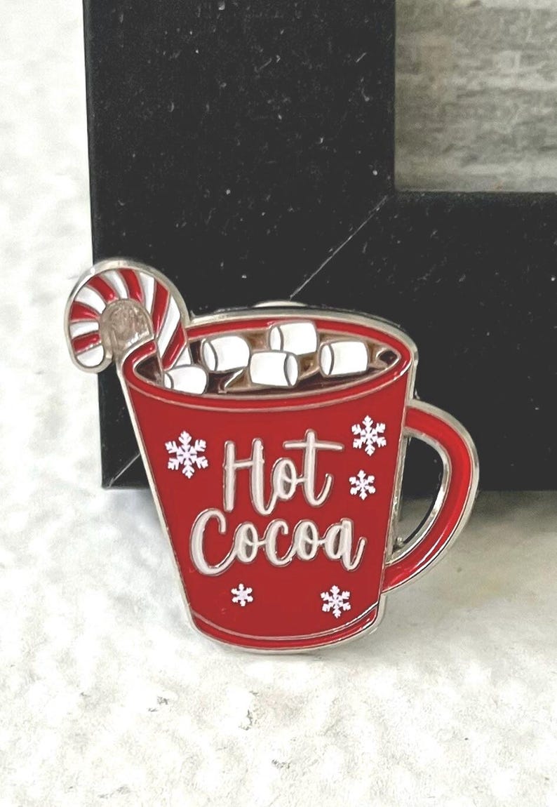 May include: Red enamel pin shaped like a mug of hot cocoa with marshmallows and a candy cane. The mug has white snowflakes and the text "Hot Cocoa" on it.