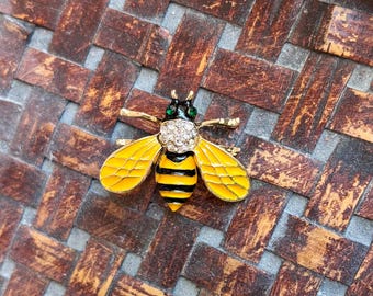 May include: A gold-toned bee brooch with yellow enamel wings and a black and yellow striped body. The bee's head is adorned with clear crystals and green accents. The brooch is a decorative accessory.