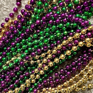 May include: Close-up of Mardi Gras beads in gold, green, and purple. The beads are round and strung together, creating a festive and colorful display. The beads are a popular accessory for celebrations.
