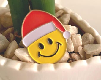 May include: A yellow smiley face pin with a red Santa hat and white trim. The pin is set against a background of white stones and green bamboo stalks, creating a festive and cheerful design.