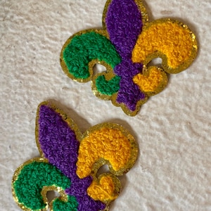 May include: Two fleur-de-lis patches in the traditional Mardi Gras colors: purple, green, and gold. The patches have a textured, embroidered appearance with gold glitter edging. The background is a textured, off-white surface.