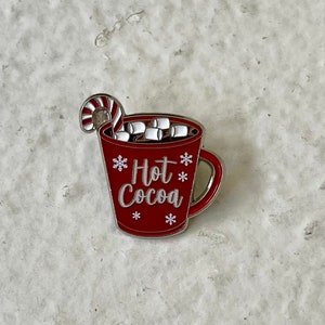 May include: Red enamel pin of a mug with a candy cane sticking out of the top. The mug is filled with white marshmallows and has the text "Hot Cocoa" on the front.