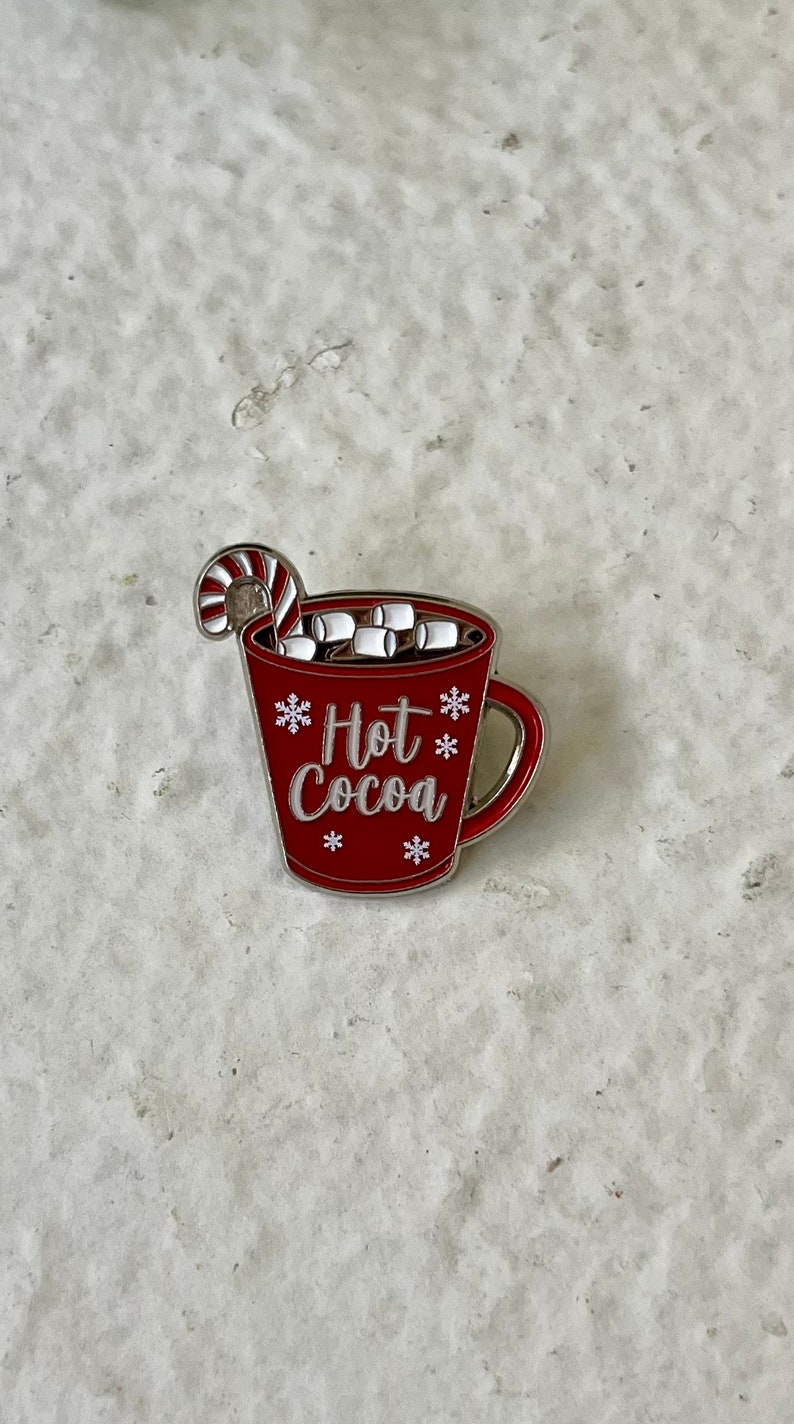 May include: Red enamel pin of a mug with a candy cane sticking out of the top. The mug is filled with white marshmallows and has the text "Hot Cocoa" on the front.