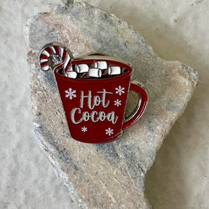 May include: Red enamel pin of a mug with a candy cane and marshmallows, with the text "Hot Cocoa" and snowflakes.