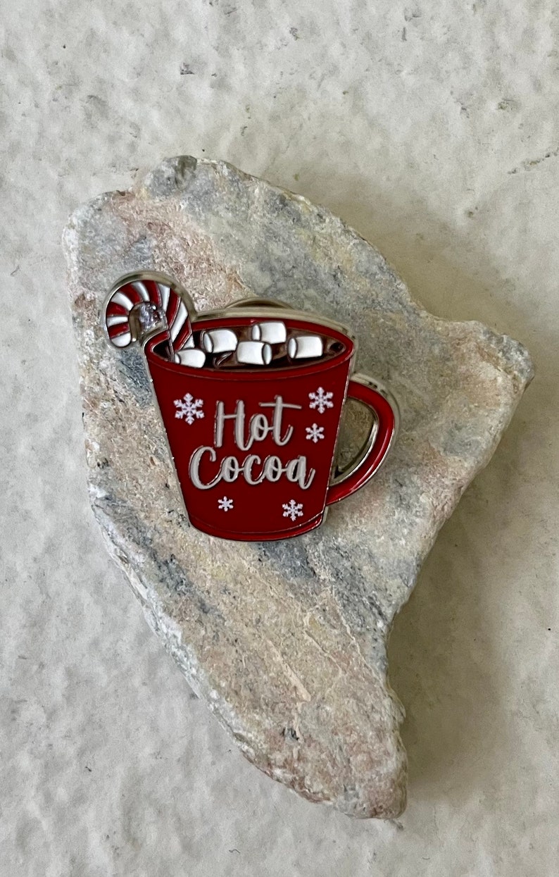 May include: Red enamel pin of a mug with a candy cane and marshmallows, with the text "Hot Cocoa" and snowflakes.
