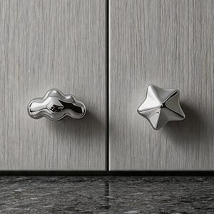 May include: Two polished silver-toned metal cabinet knobs mounted on light gray wood-grain cabinet doors. One knob is shaped like a rounded, puffy cloud, and the other knob is shaped like a five-pointed star with smooth, reflective surfaces.