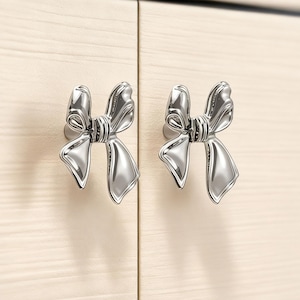 Könnte beinhalten: Zwei silberfarbene, schleifenförmige Schrankknöpfe, die an einem hellen Holzschrank befestigt sind. Die Knöpfe haben ein detailliertes Design, wobei die Schlaufen und der Knoten des Bogens deutlich sichtbar sind. Die Schranktüren haben ein helles Holzmaserungsmuster.