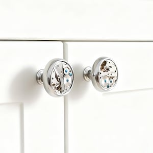 May include: Two silver cabinet knobs with watch movement faces. The knobs are round with a polished finish, and the watch mechanisms feature small blue accents. They are mounted on a white cabinet door.