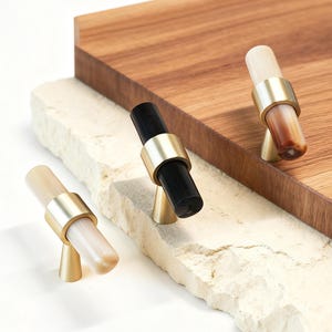 Peut inclure: Trois boutons de meuble avec des poignées cylindriques en bois clair, noir et marbré marron et crème. Chaque bouton a une bande métallique dorée et une base conique. Les boutons sont présentés sur une surface en pierre blanche avec une planche à découper en bois en arrière-plan.