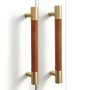 Peut inclure: Deux poignées de meuble de conception moderne. Les poignées ont une forme cylindrique avec un centre marron semblable à du cuir et des extrémités en métal doré. Elles sont montées sur une surface blanche.