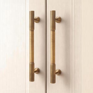 Puede incluir: Dos tiradores de armario en tono latón con un diseño de estrías verticales. Los tiradores están montados en un armario de madera de color claro. El herraje tiene forma cilíndrica con agarre texturizado y placas de montaje redondas.