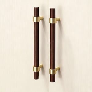 May include: Two cabinet pulls with dark brown wooden handles and gold-colored metal accents. The pulls are cylindrical and mounted on a light-colored cabinet door. The design is modern and minimalist.