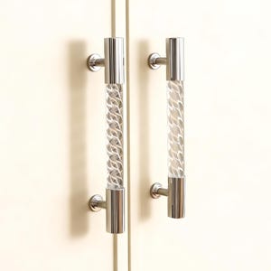 May include: Two cabinet door handles with a clear, textured center and polished chrome ends. The handles are cylindrical with a decorative pattern inside the clear center. They are mounted on a light-colored cabinet door.