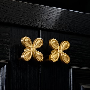 May include: Two gold-coloured, flower-shaped cabinet knobs are mounted on a black wooden cabinet door. The knobs have a four-petal design with a square centre. The wood grain is visible.
