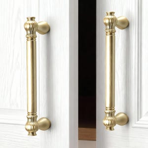 Puede incluir: Dos tiradores dorados para armarios con un diseño de barra vertical y extremos decorativos. Los tiradores están montados en puertas de armario blancas con textura de vetas de madera. Los tiradores son de color oro cálido.