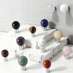 May include: A collection of decorative cabinet knobs with spherical gemstone designs. The knobs feature various colors and patterns, including black, blue, tiger's eye, amethyst, marble, and rose quartz. Each knob is mounted on a silver base.