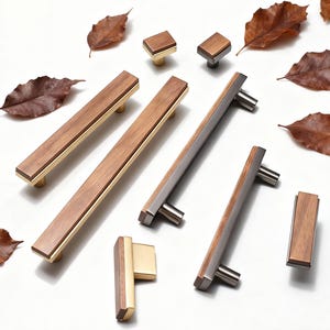 Könnte beinhalten: Eine Auswahl an Holzschrankgriffen und -knöpfen. Die Griffe haben einen rechteckigen Holzteil mit goldenen oder silbernen Metalldetails. Die Knöpfe haben eine quadratische Holzoberseite und einen Metallsockel. Braune Blätter sind um die Hardware angeordnet.