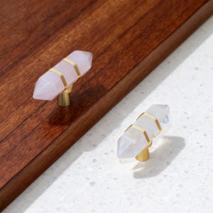 May include: Two cabinet knobs featuring double-terminated crystal points wrapped in thin gold-toned wire. One knob is pale pink, and the other is translucent white. Each crystal is mounted on a small, cylindrical gold-toned metal base.