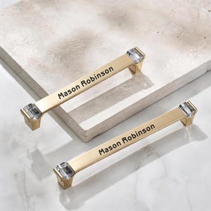 May include: Two gold-toned metal drawer pulls featuring clear, faceted crystal accents at each end. The center of each handle is engraved with the name Mason Robinson in a clean, black sans-serif font, providing a clear and legible appearance.