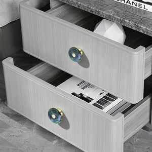 May include: A light gray wood-grain nightstand with two drawers, each featuring a round knob with a blue marbled pattern and a gold center. The top drawer is partially open, revealing a white geometric object and a book titled CHANEL.