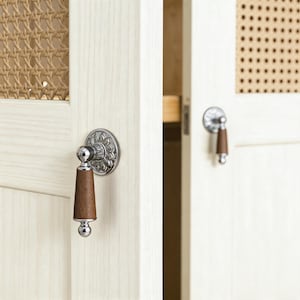 May include: A cabinet door featuring a light wood frame with a woven cane panel insert. The door is equipped with a drop-style handle consisting of a polished chrome circular base with a floral pattern and a tapered dark brown wooden grip.
