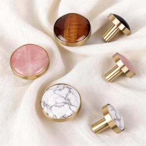 May include: A selection of cabinet knobs with different stone and marble patterns. The knobs have a gold-coloured metal base and include rose quartz, tiger's eye, marble, and black stone options. The knobs are round and have a simple, elegant design.