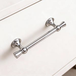 May include: A polished chrome drawer pull with a bar handle and decorative end pieces. The hardware is mounted on a white surface, showcasing its reflective quality and classic design. This cabinet hardware is suitable for various furniture styles.