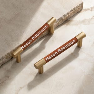 May include: Two cylindrical cabinet pulls featuring a brushed gold metal finish on the ends and a brown leather-like centre section. The text Mason Robinson is printed in white on the brown section of each handle, which has two mounting posts.