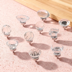 Puede incluir: Una colección de pomos de cristal transparente para armarios en varias formas, incluyendo diseños redondos, ovalados y facetados. Cada pomo tiene una base plateada. Los pomos están dispuestos sobre una superficie rosa claro, con una tabla de cortar de madera en el fondo.