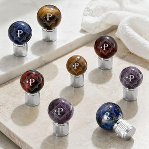 May include: A collection of eight spherical bottle stoppers featuring polished gemstones in various colours including blue, brown, red, and purple. Each stone is engraved with the letter P inside a geometric frame and attached to a silver base.