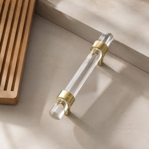 May include: A cabinet pull featuring a clear, cylindrical acrylic handle accented by two brushed gold metal mounting posts. The hardware has a sleek, linear design with circular end caps and a polished finish on the transparent center section.