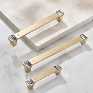 May include: Three gold-toned metal cabinet pulls of varying lengths, each featuring a brushed finish and clear, faceted crystal accents at both ends. Each handle has a small, engraved logo detail near the crystal on the top surface.