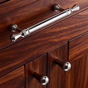 May include: Close-up of a wooden cabinet with a polished, dark brown finish. The cabinet features a long, silver handle and two smaller, round knobs with silver accents. The wood grain is visible.