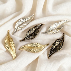 Puede incluir: Un conjunto de seis tiradores u ornamentos decorativos de metal con forma de hoja en varios acabados, incluyendo plata pulida, oro brillante, bronce oscuro y latón antiguo, con venas en relieve detalladas y una forma alargada.