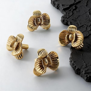 May include: Four gold-coloured, flower-shaped cabinet knobs. Each knob features a detailed floral design with multiple petals and a central stamen. The knobs are made of a shiny, metallic material.