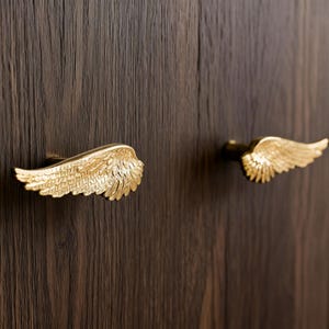 May include: Two gold-coloured, wing-shaped cabinet pulls are mounted on a dark brown wooden surface. The pulls are detailed with feather-like textures, adding a decorative touch to the cabinetry. The hardware is designed to enhance the aesthetic of furniture.