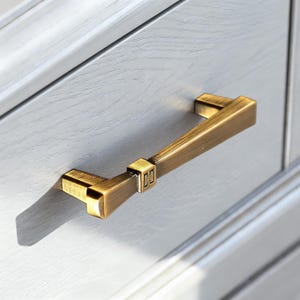Può includere: Una maniglia per cassetto in metallo color ottone con finitura spazzolata e un dettaglio quadrato centrale che riporta le lettere CJ. La maniglia è montata orizzontalmente su un frontale di cassetto in legno grigio chiaro con venature.