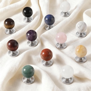 May include: A selection of decorative cabinet knobs with spherical gemstone tops in various colours, including black, purple, pink, and yellow. Each knob has a silver-coloured, spiral base, and they are arranged on a white fabric surface.