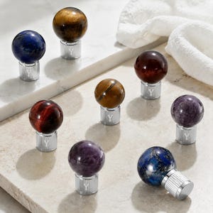 May include: A collection of eight spherical gemstone knobs, each featuring a polished stone ball in various colors including blue, brown, red, and purple, mounted on a cylindrical silver-toned metal base with a vertical ribbed texture and a letter Y.