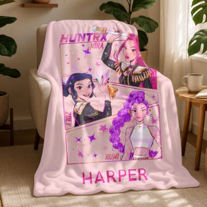 Op de afbeelding: Een lichtroze deken met een kleurrijke afbeelding van drie vrouwen in anime-stijl. De tekst "HUNTX MIRA", "ZOEY", "RUMI" en "HARPER" is aanwezig. De deken is over een beige fauteuil gedrapeerd.