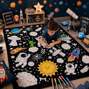 Op de afbeelding: Een zwart rechthoekig kleed met een wit lijntekening-thema van de ruimte, inclusief planeten, sterren, een astronaut, een raket en een UFO. Het kleed bevat een grote gele zon met een lachend gezicht en wordt ingekleurd met stiften.